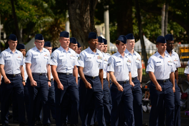 Männer in Uniform auf einer Straße mit einem Eisengeländer dahinter, Bäume und Pfähle im Hintergrund.