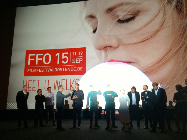 Eine Gruppe von Menschen auf einer Bühne mit einem Mann in der Mitte, der ein Mikrofon hält, ein Display hinter ihnen mit dem Text "Foto Filmfestivaloostende 2015" und einem Bild einer Frau, sowie einigen Menschen neben dem Display.