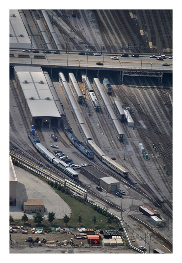 Eine Brücke mit wenigen Fahrzeugen darauf, darunter Eisenbahnschienen mit wenigen Zügen und eine Landschaft mit Gebäuden, Bäumen, Masten und diversen Gegenständen und Fahrzeugen am Boden.