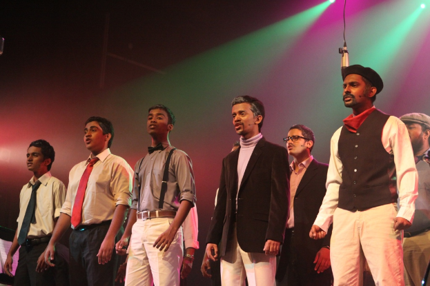 Eine Gruppe von Männern steht auf einer Bühne und führt ein Drama auf, während grüne Scheinwerfer hinter ihnen leuchten.