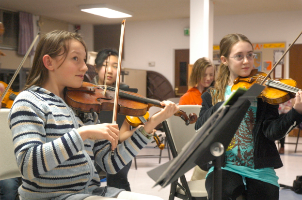 Eine Gruppe von Frauen, die auf Stühlen sitzen und Geigen spielen, mit einem Buch auf einem Ständer, einer Tür, einer Säule, einem Vorhang und einem Fenster im Hintergrund.