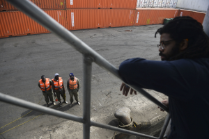 Ein Mann in legerer Kleidung steht auf einem Metallgeländer in der Nähe einer Gruppe von Menschen in orangefarbenen Jacken und Kappen, mit Containern und einer Wand im Hintergrund am Hafen von Los Angeles.