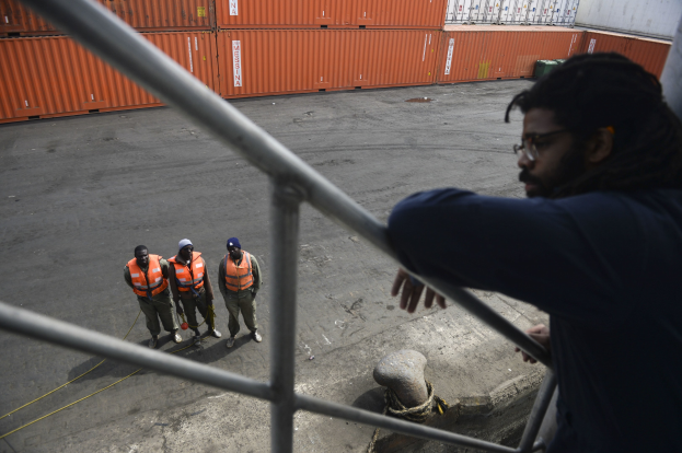 Ein Mann in legerer Kleidung steht auf einem Metallgeländer in der Nähe einer Gruppe von Menschen in orangefarbenen Jacken und Kappen, mit Containern und einer Wand im Hintergrund am Hafen von Los Angeles.