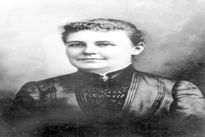 Schwarzes und weißes Foto einer Frau in einem Kleid, die mit gefalteten Händen und erhobenem Kopf steht und Entschlossenheit und Stärke ausstrahlt.