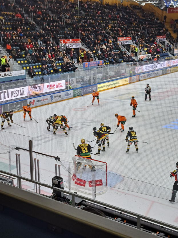 Eine Gruppe von Menschen spielt Hockey auf einem Eisstadion mit einem Netz und einer Umzäunung, Zuschauern im Hintergrund und Bannern mit Text, unter Stadionbeleuchtung.