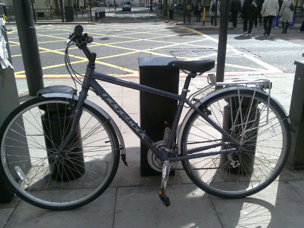 Ein Fahrrad steht im Vordergrund mit Metallstangen und Pfosten dahinter, ein Fahrzeug auf der Stra√üe, Passanten, die gehen, Metallstabz√§une, M√ľlltonnen, Ampeln und Geb√§ude im Hintergrund.