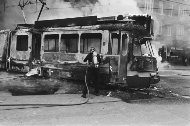 Schwarzes und wei├čes Bild eines Mannes in einem Helm, der eine Pfeife h├Ąlt und neben einer ausgebrannten Stra├čenbahn auf der Stra├če steht, mit Rauch aufsteigend, ├ťberkopfleitungen und ein Geb├Ąude im Hintergrund.