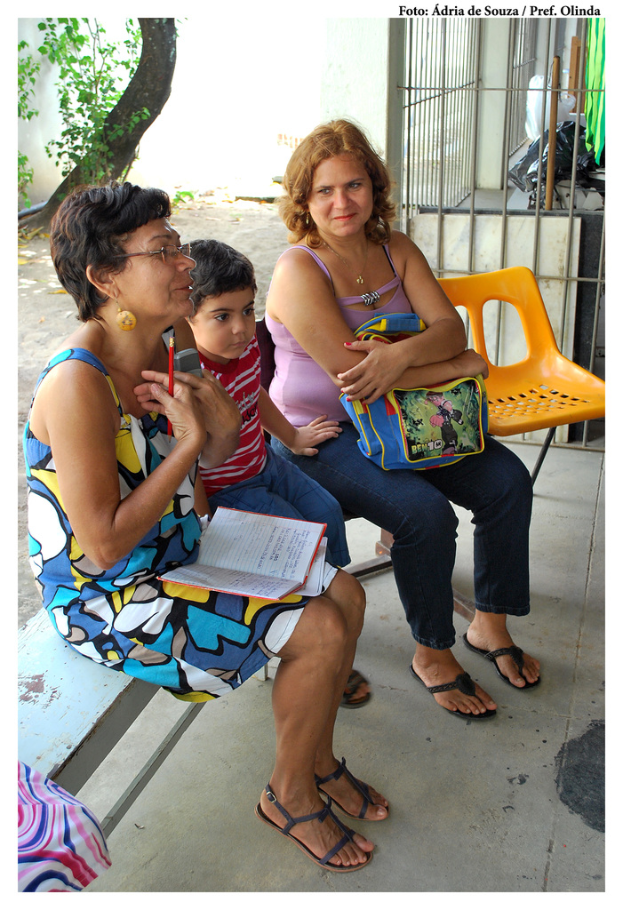 Zwei Frauen und ein Kind auf einer Bank sitzend mit einem orangefarbenen Stuhl daneben, einem Baum und Pflanzen im Hintergrund.