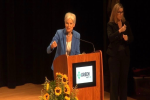 Zwei Frauen stehen an einem Podium mit einem Mikrofon, einem Bildschirm und einem Blumenstrauß vor einem dunklen Hintergrund auf einer Grünen Veranstaltung.