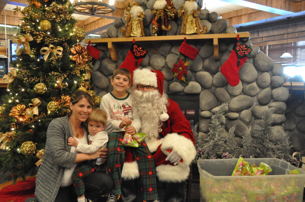 Eine Frau, Kinder und ein Mann als Santa Claus mit Brille und Handschuhen stehen neben einem geschmückten Weihnachtsbaum, einer Wand mit Steinen, Spielzeugen, Dekorationen, einem Korb mit Gegenständen und Lichtern im Hintergrund. Ein Junge hält ein Päckchen.