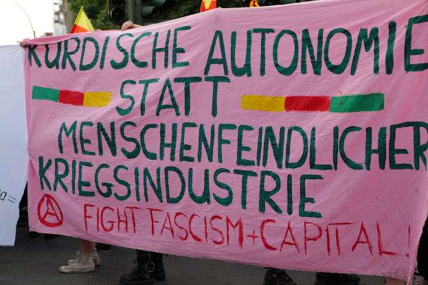 Eine Gruppe von Menschen steht auf einer Straße und hält ein pinkes Banner hoch, auf dem "Kurdische Autonomie Statt Menschenfeindlicher Kriegsindustrie Fight Fascism Capital" steht, mit Verkehrszeichen, Fahnen, Bäumen, Gebäuden und einem klaren blauen Himmel im Hintergrund.