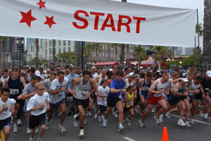 Gruppe von Menschen, die bei einem Marathon laufen, mit einem Verkehrskegel im Vordergrund und einem Banner im Hintergrund, umgeben von Bäumen, Laternenmasten, Gebäuden und einem klaren blauen Himmel.