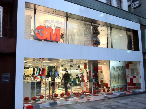 Ein Gebäude mit einer Glaswand, auf der Kleider an Bügeln, eine Schaufensterpuppe, ein Fahrrad, aufgeklebte Bilder und Schrift auf der Scheibe zu sehen sind.