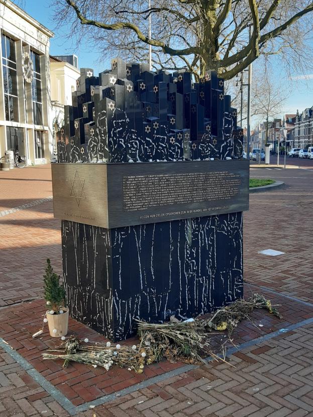 Denkmal in einem Stadtplatz für Holocaust-Opfer, mit einem Baum davor, einem Topfgewächs auf dem Boden, verstreuten trockenen Blättern und Gebäuden, Fahrzeugen und Bäumen im Hintergrund.