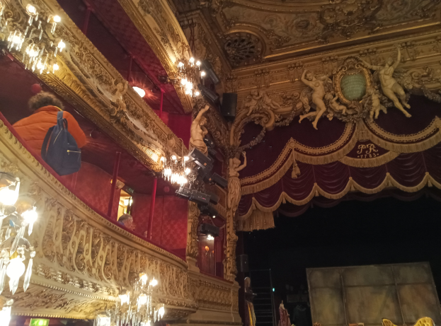 Das Innere eines Theaters mit Kronleuchtern an der Decke, Skulpturen an den Wänden und Menschen auf der Bühne, mit verstreuten Gegenständen, die eine lebhafte Atmosphäre schaffen.