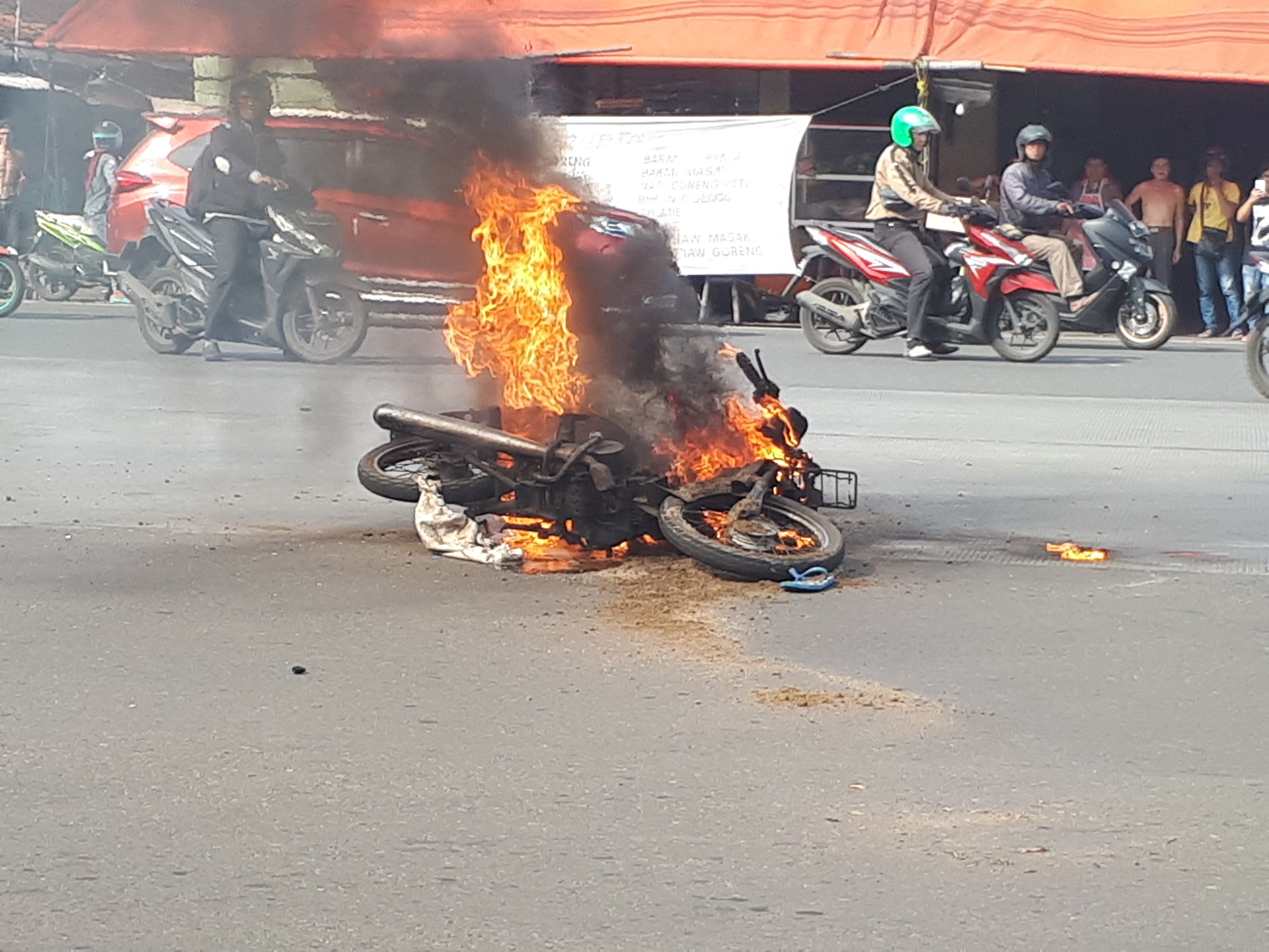 Ein in Flammen stehendes Motorrad am Straßenrand, mit einem Auto und einem Van im Hintergrund, umgeben von Menschen, einige tragen Helme, eine Fahne mit Text und ein Zelt.