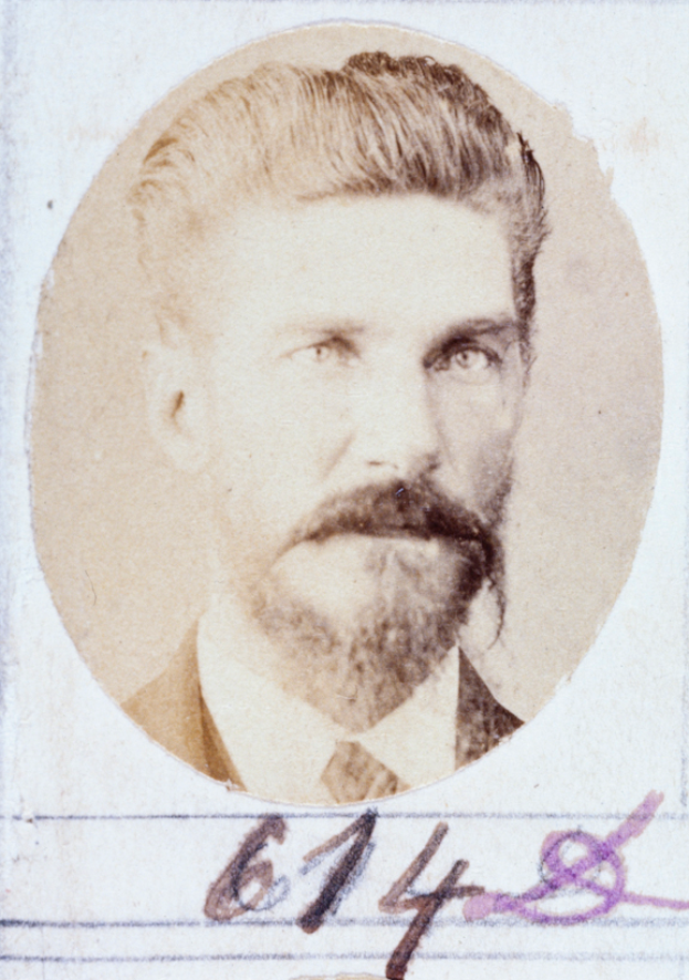 Ein altes Foto eines Mannes mit Bart und Schnurrbart, der direkt in die Kamera schaut und leicht geöffnete Lippen hat, ordentlich gekämmtes Haar und einen gut gepflegten Bart.