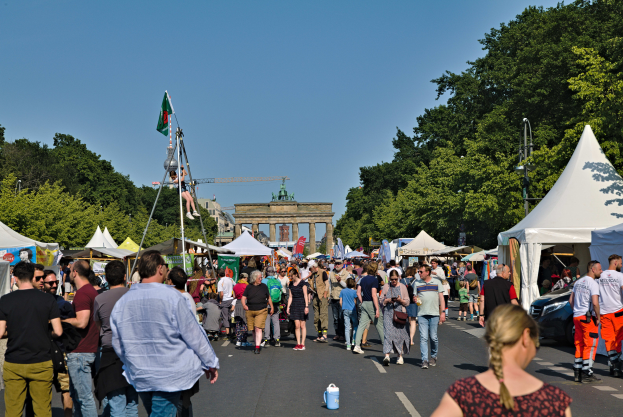 Eine Menschenmenge geht eine Straße entlang, gesäumt von Zelten, Fahrzeugen und Bäumen, mit einem Bogen und einem klaren blauen Himmel im Hintergrund und Polen mit Fahnen auf der linken Seite, wahrscheinlich das Oktoberfest in München, Deutschland.