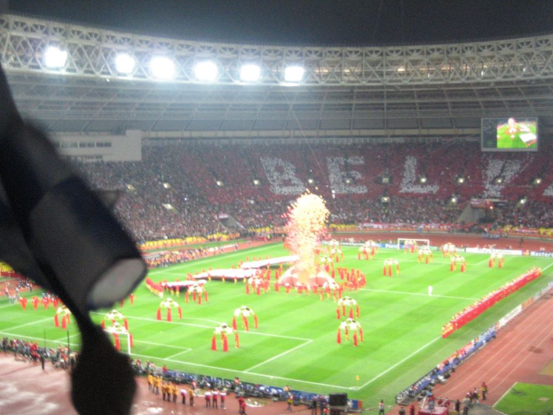 Ein Stadion mit Flutlicht, Eisengittern, Zuschauern, stehenden Personen, Werbetafeln, Anzeigetafeln, Sicherheitskameras, Fahnen und Deko.