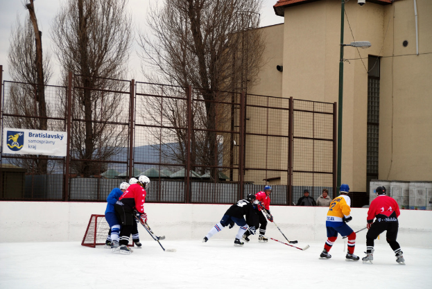 Personen spielen Eishockey auf einem Eisstadion mit Gebäuden, Bäumen, einer Straßenlaterne, einem Namensschild und Zäunen im Hintergrund bei klarem Himmel.