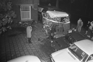 Ein Schwarz-Weiß-Foto eines verbeulten Autos am Straßenrand, umgeben von anderen Autos, Menschen, Fahrrädern, Bäumen und einem Gebäude.