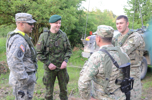 Vier Soldaten in Uniformen und Mützen stehen und sprechen, mit einem Fahrzeug auf der rechten Seite und Bäumen im Hintergrund.