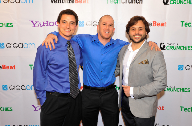 Drei Personen vor einer Sponsoren-Plakate stehend, bekleidet.