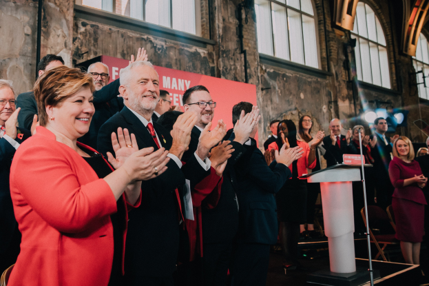 Eine Gruppe von Menschen, die vor einem Publikum applaudieren, mit einem Podium, einem Mikrofon und einer Tafel mit Text auf der rechten Seite und Stühlen, einer Fahne, einer Wand, Fenstern und Lichtern im Hintergrund.