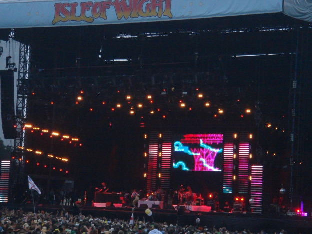 Eine B√ľhne mit Lichtern, einem Bildschirm und Musikern, die Instrumente spielen, mit einem Banner oben, einer Menge unten und Fahnen mit Stangen und Ballons drumherum.