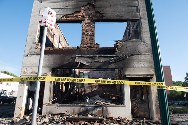 Ein abgerissener Bau inmitten einer Straße, umgeben von gelbem Absperrband, mit Trümmern drumherum, anderen Gebäuden, Fahrzeugen, Bäumen und einem klaren blauen Himmel im Hintergrund.