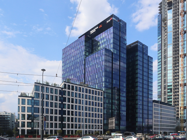 Großes modernes Bürogebäude mit vielen Fenstern, identifiziert als Deutsche Bank Hauptsitz in Frankfurt, Deutschland, umgeben von städtischer Infrastruktur einschließlich Straßenlaternen, Strommasten, Fahrzeugen, Grünflächen und einem bewölkten Himmel.