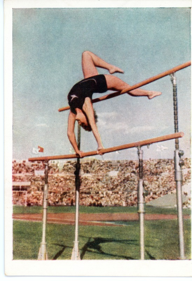 Eine Frau führt einen Hochsprung an parallelen Stangen in einem Stadion mit Zuschauern unter einem klaren blauen Himmel durch.