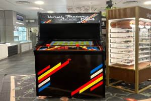 Ein Geschäftsinnenraum mit einem zentralen Retro-Arcade-Automaten, einer Glasvitrine auf der rechten Seite und einer retro-stylischen Beleuchtung mit Schränken, einem Fernseher und anderen Gegenständen im Hintergrund.