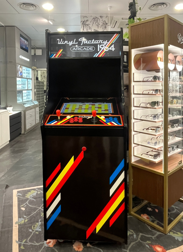 Ein Geschäftsinnenraum mit einem zentralen Retro-Arcade-Automaten, einer Glasvitrine auf der rechten Seite und einer retro-stylischen Beleuchtung mit Schränken, einem Fernseher und anderen Gegenständen im Hintergrund.