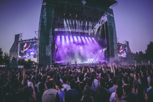 Eine große Menschenmenge auf einem Musikfestival schaut auf einer beleuchteten Bühne mit Instrumenten und Bildschirmen, die vor einem Gebäude, Bäumen und einem klaren blauen Himmel aufgebaut sind.