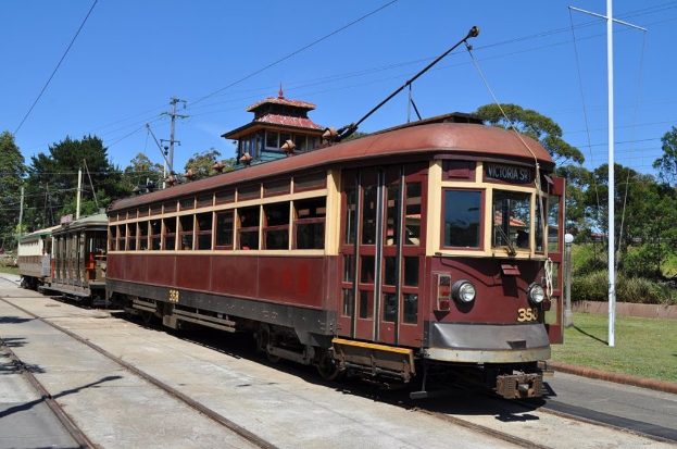 Ein Zug auf Schienen mit Masten, Kabeln, Bäumen und Häusern im Hintergrund.