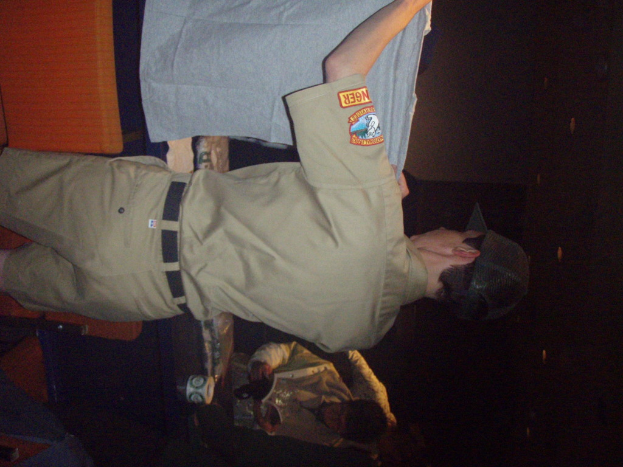 Ein Polizeibeamter hält ein T-Shirt in den Vordergrund, im Hintergrund ein Junge mit einem Buch und einem Becher, der neben einem Schreibtisch und Lampen steht.