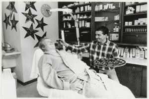 Ein Mann lächelt in einem Zahnarztstuhl, während ein anderer Mann neben ihm steht und ein Tablett hält, mit einem Waschbecken, Uhr, Sternen an der Wand und Regalen voller Flaschen im Hintergrund.