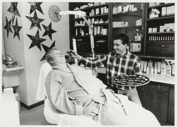 Ein Mann lächelt in einem Zahnarztstuhl, während ein anderer Mann neben ihm steht und ein Tablett hält, mit einem Waschbecken, Uhr, Sternen an der Wand und Regalen voller Flaschen im Hintergrund.