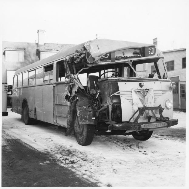 Schwarzes und weißes Foto eines schwer beschädigten Busses mit eingedellter Front und zersplitterten Fenstern an der Seite einer Straße, mit Gebäuden im Hintergrund.