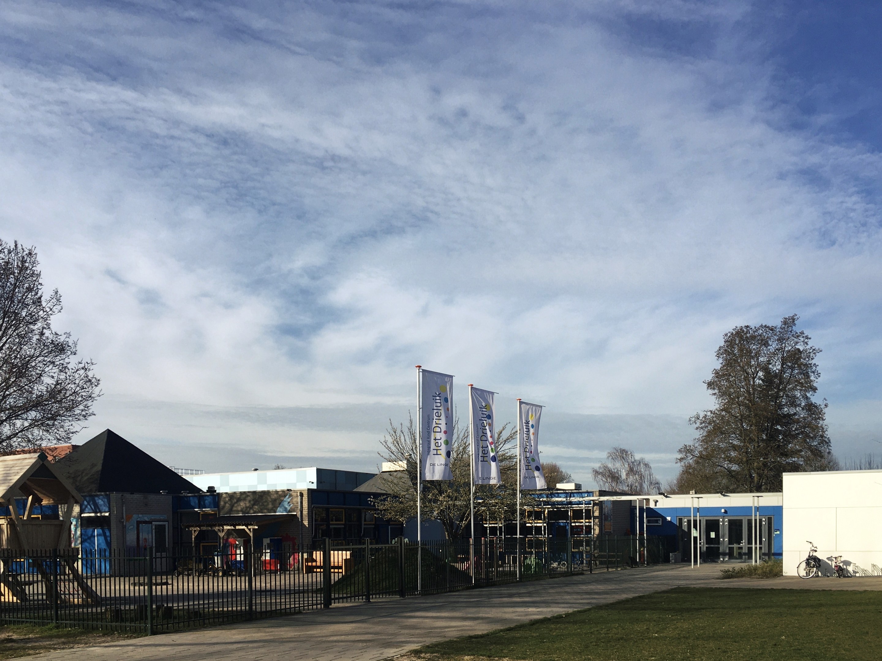 Großes Gebäude mit Metallzaun, Rasenfläche, Weg, Bäumen, Pfosten, Bannern, einem geparkten Fahrrad, nahen Häusern und einem bewölkten Himmel, identifiziert als der neue Spielplatz der Schule.