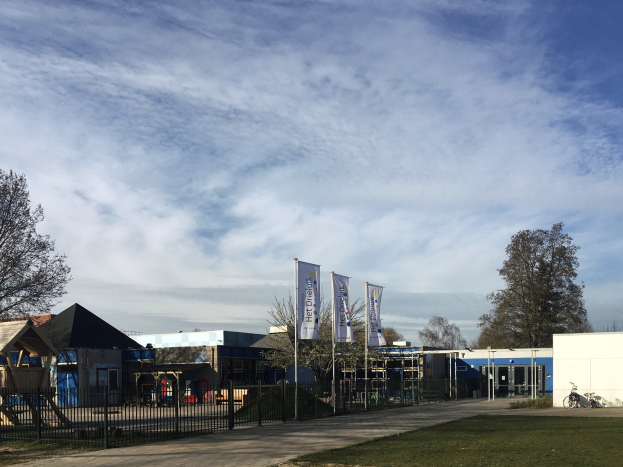 Großes Gebäude mit Metallzaun, Rasenfläche, Weg, Bäumen, Pfosten, Bannern, einem geparkten Fahrrad, nahen Häusern und einem bewölkten Himmel, identifiziert als der neue Spielplatz der Schule.