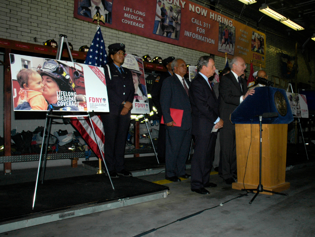 Eine Gruppe von Menschen steht neben einem Podium mit Mikrofonen, Schildern auf Ständern, Fahnen und Schildern an der Wand, alles unter hellen Lichtern.