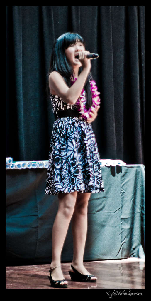 Eine Frau in einem bunten Kleid singt in ein Mikrofon auf der B├╝hne, mit einem tisch voller Papiere und einem Vorhang im Hintergrund.