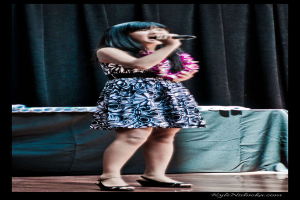 Eine Frau in einem bunten Kleid singt in ein Mikrofon auf der B├╝hne, mit einem tisch voller Papiere und einem Vorhang im Hintergrund.