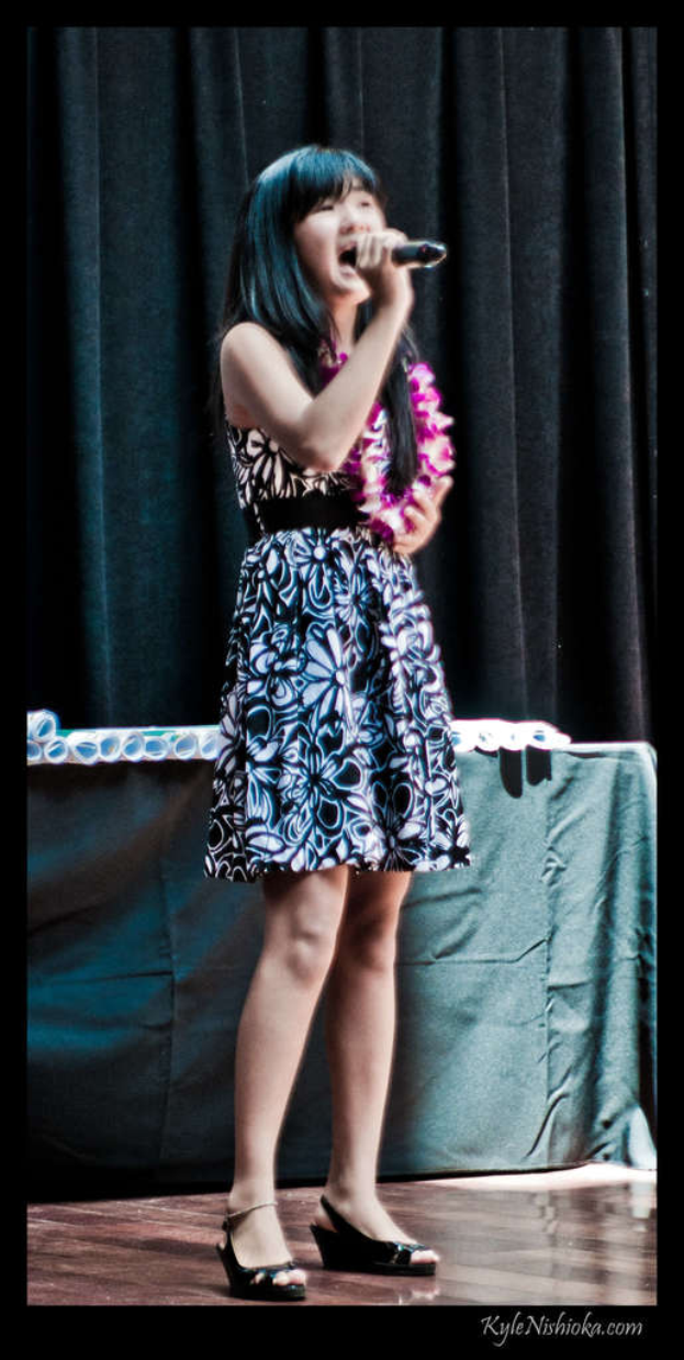 Eine Frau in einem bunten Kleid singt in ein Mikrofon auf der B├╝hne, mit einem tisch voller Papiere und einem Vorhang im Hintergrund.