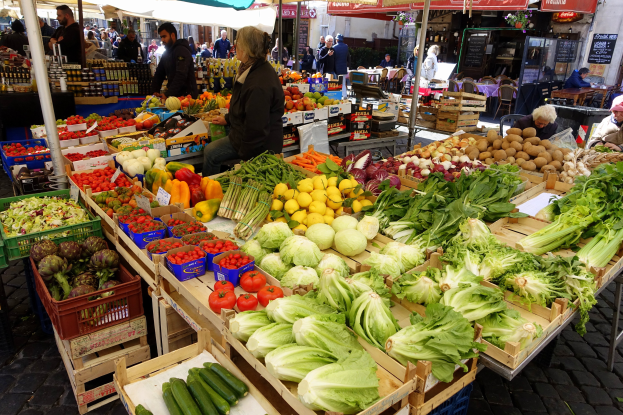 Ein belebter Outdoor-Markt mit verschiedenen Früchten und Gemüsen auf einem Tisch, Menschen, die umhergehen, einige sitzen und andere stehen, unter Zelten mit Gebäuden und Texttafeln im Hintergrund.