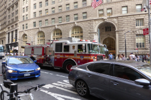 Ein Feuerwehrauto steht an der Seite einer Straße in New York City, mit Fahrzeugen auf der Straße, Fußgängern auf dem Gehweg und Fahrrädern in der Nähe, mit Gebäuden, Laternenmasten und Fahnenmasten im Hintergrund.