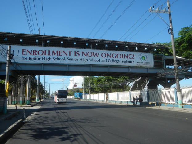 Ein Bus fährt unter einer Brücke mit einem "Jetzt Anmelden"-Schild, während Fußgänger darunter gehen und Strommasten die Straße mit Bäumen und einem blauen Himmel im Hintergrund säumen.