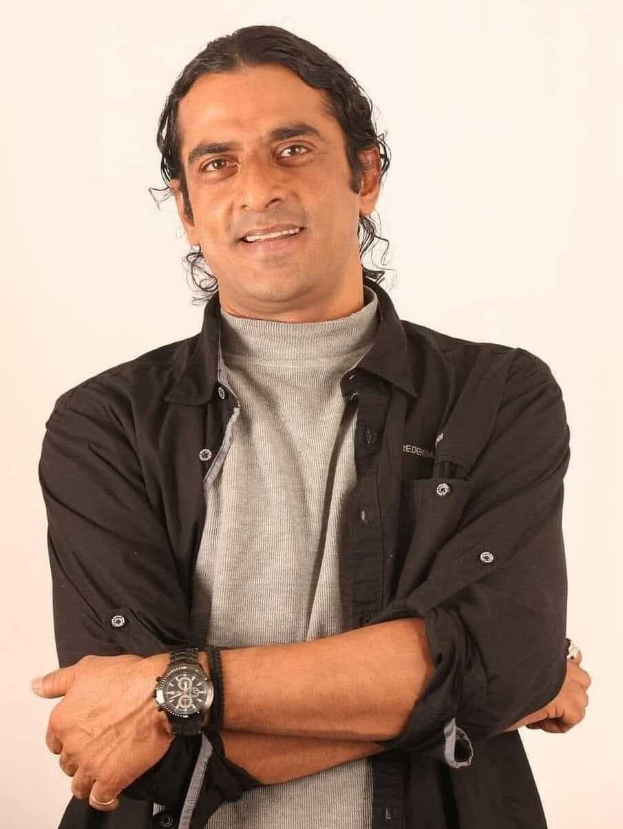 Ein Mann in einem schwarzen und aschfarbenen Kleid mit überschlagenen Armen, der eine Uhr tr├Ągt, vor einem wei├čen Hintergrund steht und nachdenklich scheint.